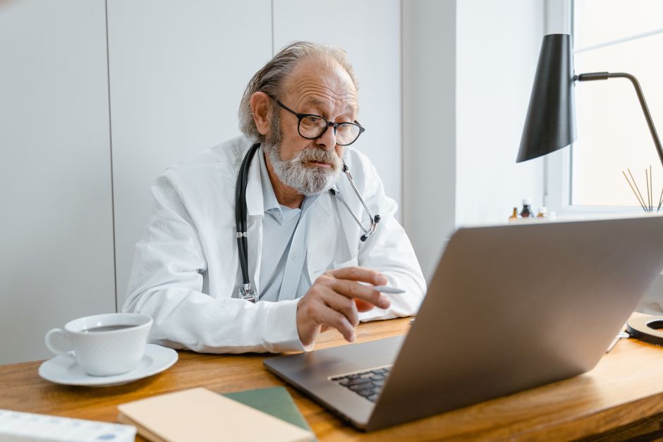 FSATA Designation Senior male doctor using laptop for telehealth consultation in office setting.