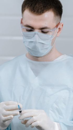 Healthcare professional wearing protective gear and preparing an injection in a medical setting.